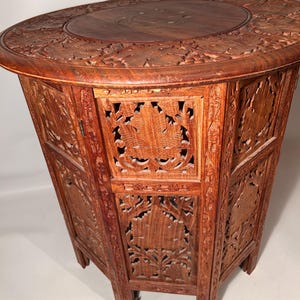 May include: A reddish-brown wooden table with detailed floral carvings. The oval tabletop and hexagonal base feature openwork panels. The craftsmanship and rich colour suggest a vintage or antique piece. The table's height is approximately 60 cm.