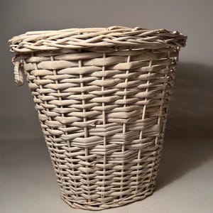 May include: A cream-coloured woven wicker basket. The basket has a tapered shape, wider at the top and narrowing towards the base. It features a woven handle on one side and a rim around the top. The basket is designed for storage.