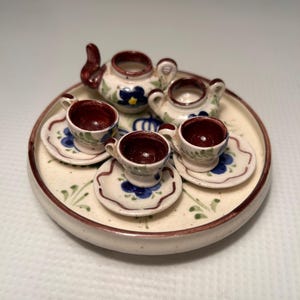 May include: A miniature ceramic tea set featuring a teapot, creamer, sugar bowl, and two cups and saucers. The set is decorated with blue floral designs and a brown rim. The set is on a round tray with a matching design.