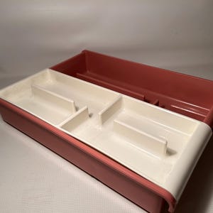 May include: A two-tone plastic organiser with a divided interior. The top section is a muted red, while the lower section is white with red trim. The organiser has several compartments for storage.