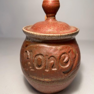 May include: A reddish-brown ceramic honey pot with a lid and a knob. The word "Honey" is embossed on the front. The pot has a rustic, textured appearance, suggesting a handmade quality.