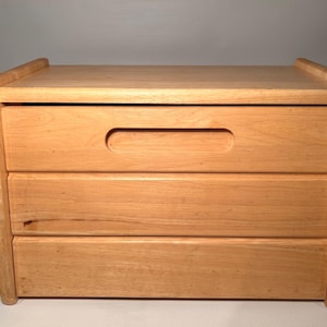 Può includere: Un comò in legno a tre cassetti con un design semplice. Il comò è in legno chiaro e ha una finitura liscia.