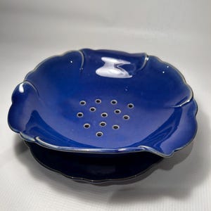 Può includere: Un colino in ceramica blu con un bordo a forma di conchiglia e un set di fori di scarico. Il colino è impilato su un piatto blu abbinato.