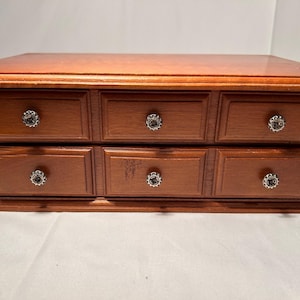 May include: A wooden jewellery box with six drawers, each with a decorative knob. The box has a rich, reddish-brown finish and a rectangular shape. The drawers are arranged in two rows of three.
