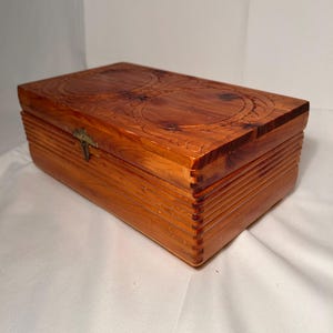 Può includere: Una scatola rettangolare in legno con una calda tonalità marrone-rossastra. Il coperchio presenta disegni circolari intagliati e la scatola ha un design a coste sui lati. Un piccolo fermaglio color ottone assicura la scatola, che sembra essere in legno di cedro.