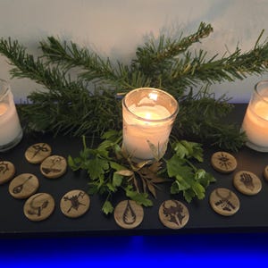 May include: A set of wooden tokens with various symbols, including a sword, a hand, a fire, and a skull. The tokens are arranged around three lit candles on a black surface. The candles are in clear glass jars and are surrounded by green foliage.