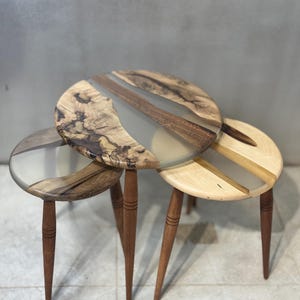May include: A trio of wooden side tables with circular tops. The tables combine natural wood and clear resin, with dark brown, light brown, and beige hues. Each table has three slender, tapered legs. The tables are approximately 40 cm in diameter.