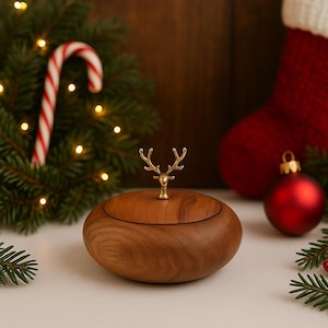 Puede incluir: Una caja de joyería de madera con una cabeza de ciervo dorada en la tapa. La caja, de forma ovalada, está sobre una superficie blanca, con decoraciones navideñas de fondo, como un calcetín y adornos rojos.