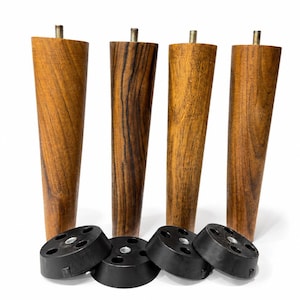 Puede incluir: Cuatro patas de mueble de madera cónicas con vetas visibles y pernos metálicos roscados en la parte superior. Cada pata se acompaña de una placa de montaje circular de plástico negro con varios orificios y un inserto metálico central.