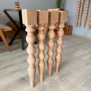 Könnte beinhalten: Vier unfertige Tischbeine aus Holz. Jedes Bein hat eine quadratische Oberseite, eine gedrehte, bauchige Mitte und einen sich verjüngenden Boden. Die Beine sind aus hellfarbigem Holz gefertigt und stehen auf einem hellfarbigen Holzboden.