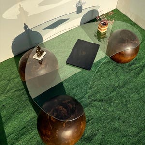 Puede incluir: Una mesa de centro de vidrio curvada con una base de madera oscura. La mesa tiene tres patas redondas de madera y una tapa de vidrio transparente. La mesa está sobre una alfombra verde.