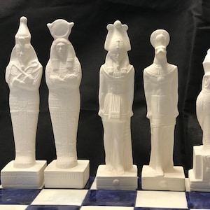 Puede incluir: Conjunto de cinco piezas de ajedrez de cerámica blanca con temática egipcia. Las piezas están en varias poses e incluyen un faraón, una reina, un dios y un halcón.