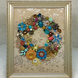 May include: A framed wall art piece featuring a circular wreath arrangement made of assorted colorful vintage jewelry, including rhinestones, beads, and metal charms, set against a beige background with a subtle floral pattern in a gold frame.