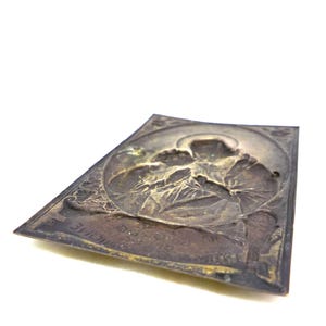 May include: A rectangular, antique metal plaque with a detailed relief of a religious figure. The plaque has a dark, aged patina with visible wear and tear. The figure is surrounded by a circular border and the plaque has text at the bottom.