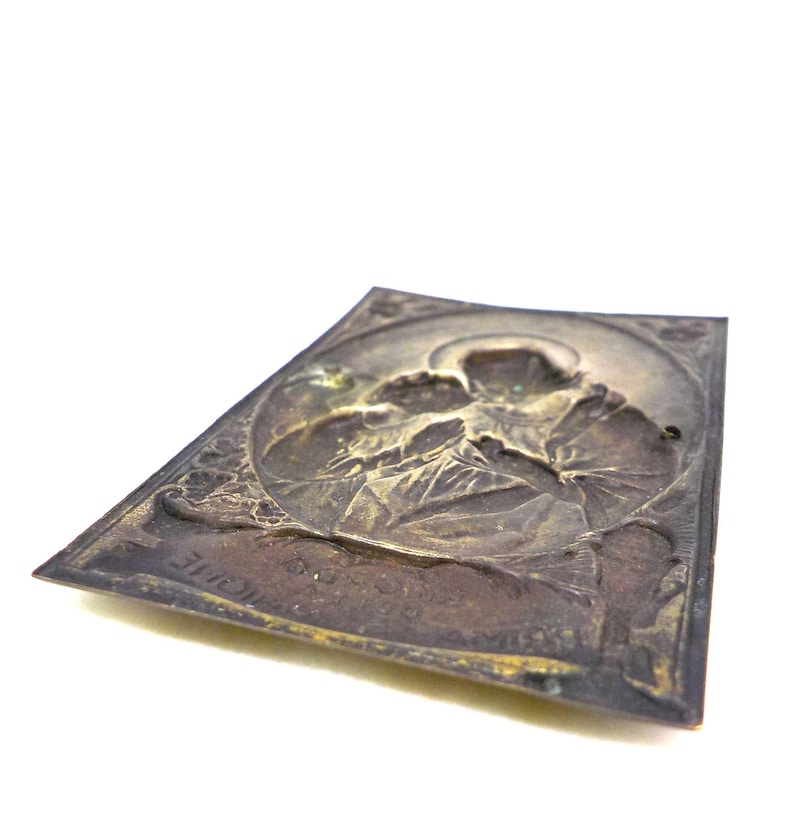 May include: A rectangular, antique metal plaque with a detailed relief of a religious figure. The plaque has a dark, aged patina with visible wear and tear. The figure is surrounded by a circular border and the plaque has text at the bottom.
