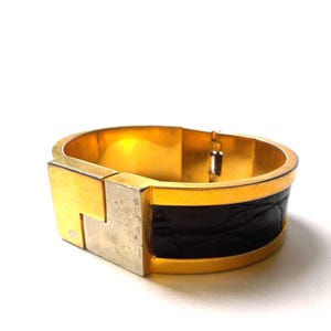 以下が含まれることがあります： 長方形の留め金と黒い革バンドが付いた、金色の金属製カフブレスレット。 ブレスレットはヒンジ開閉式で、磨かれた仕上げで、質感と色を組み合わせています。