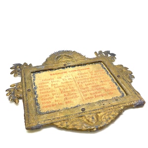 May include: An antique gold-colored frame with ornate detailing and a rectangular center. The center features a printed text in Latin, with the words "SACRUM CONVIVIUM" at the top. The frame has a weathered appearance.
