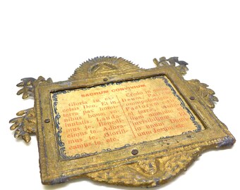 May include: An antique gold-colored frame with ornate detailing and a rectangular center. The center features a printed text in Latin, with the words "SACRUM CONVIVIUM" at the top. The frame has a weathered appearance.