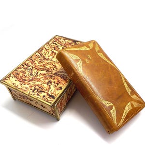 Könnte beinhalten: Eine rechteckige Schachtel mit einem marmorierten Muster in Braun- und Cremetönen, neben einem braunen, ledergebundenen Buch mit goldenen Akzenten. Die Schachtel hat eine goldene Umrandung, und das Buch hat goldene Buchstaben.