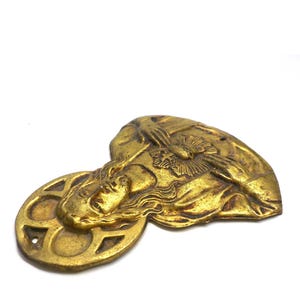 May include: A gold-colored metal religious emblem featuring a detailed relief of a person's face with flowing hair and a halo. The emblem has a circular element with geometric cutouts and a heart-shaped element.