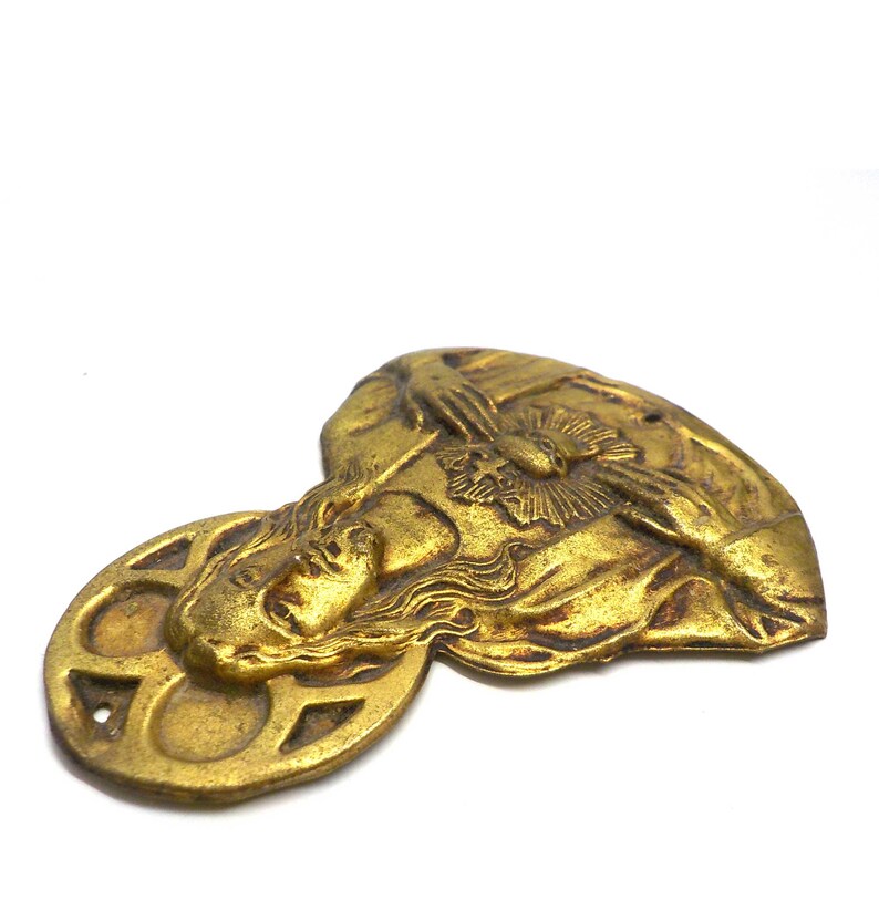 May include: A gold-colored metal religious emblem featuring a detailed relief of a person's face with flowing hair and a halo. The emblem has a circular element with geometric cutouts and a heart-shaped element.