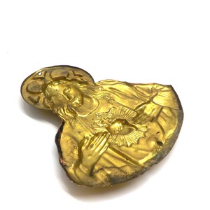May include: A vintage gold-toned religious pendant depicting a figure with a halo, hands clasped in prayer, and a heart radiating light. The pendant has a detailed relief design and a slightly worn appearance.