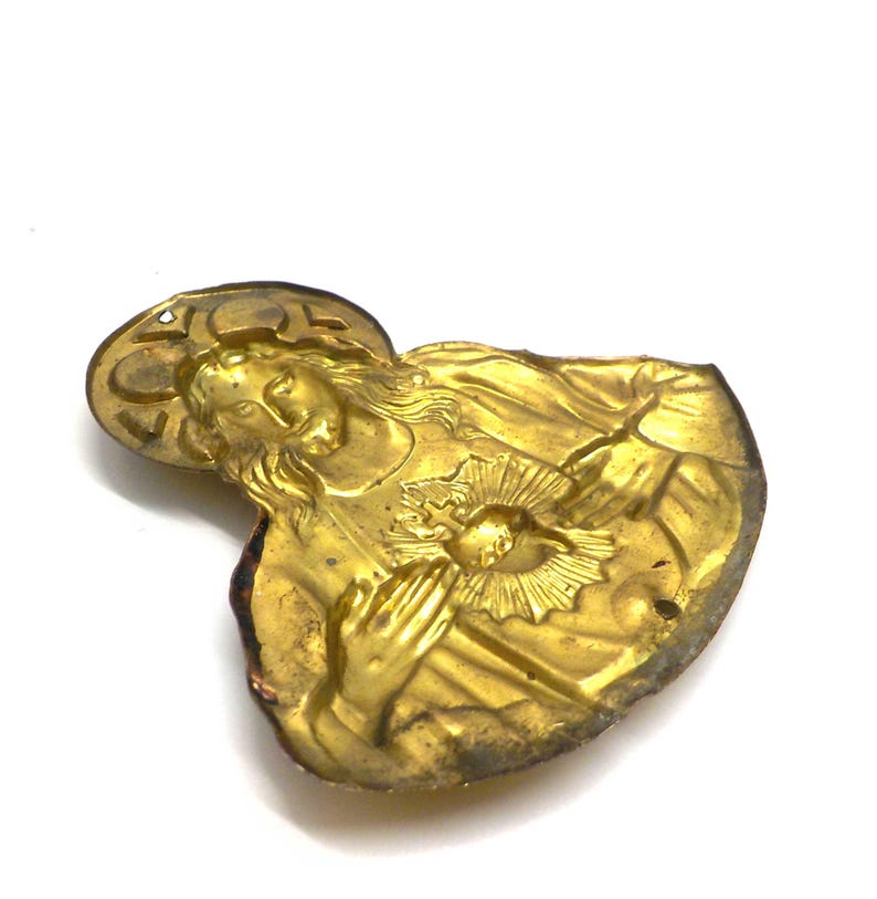 May include: A vintage gold-toned religious pendant depicting a figure with a halo, hands clasped in prayer, and a heart radiating light. The pendant has a detailed relief design and a slightly worn appearance.