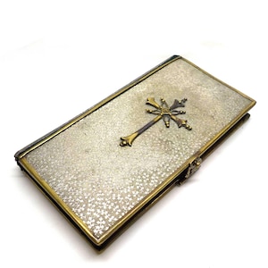 May include: A rectangular silver-toned box with a gold-coloured cross emblem. The box has a textured surface and gold-coloured trim. The cross is detailed with ornate designs. The box is closed with a small clasp.