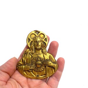 May include: A gold-colored religious pendant depicting a figure with long hair, a beard, and a halo. The figure has a heart on their chest. The pendant is held in a hand against a white background.
