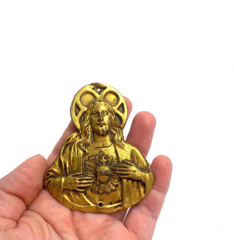 May include: A gold-colored religious pendant depicting a figure with long hair, a beard, and a halo. The figure has a heart on their chest. The pendant is held in a hand against a white background.
