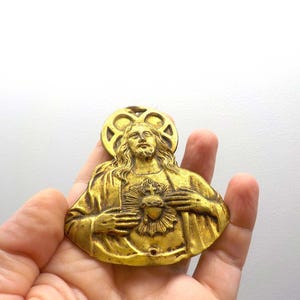 May include: A vintage gold-toned religious pendant depicting a figure with long hair, a halo, and a heart. The pendant is held in a hand against a white background. The figure is wearing a robe and has a serene expression.