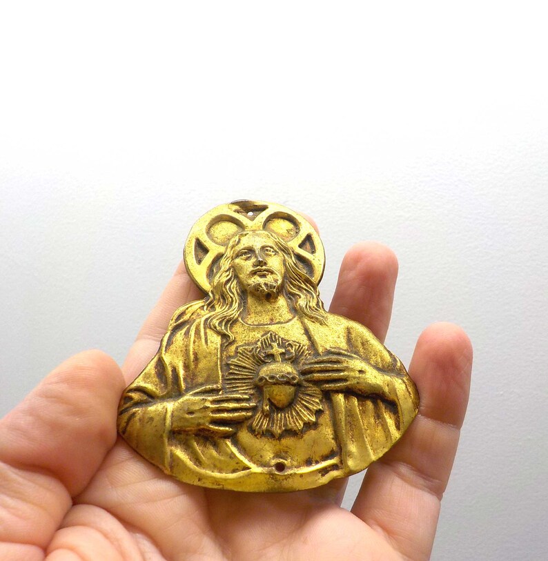 May include: A vintage gold-toned religious pendant depicting a figure with long hair, a halo, and a heart. The pendant is held in a hand against a white background. The figure is wearing a robe and has a serene expression.