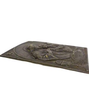 May include: A bronze-toned rectangular plaque with a relief of a religious figure. The figure is wearing a robe and has a halo. The plaque has text along the right edge. The plaque is slightly angled, showing its thickness.