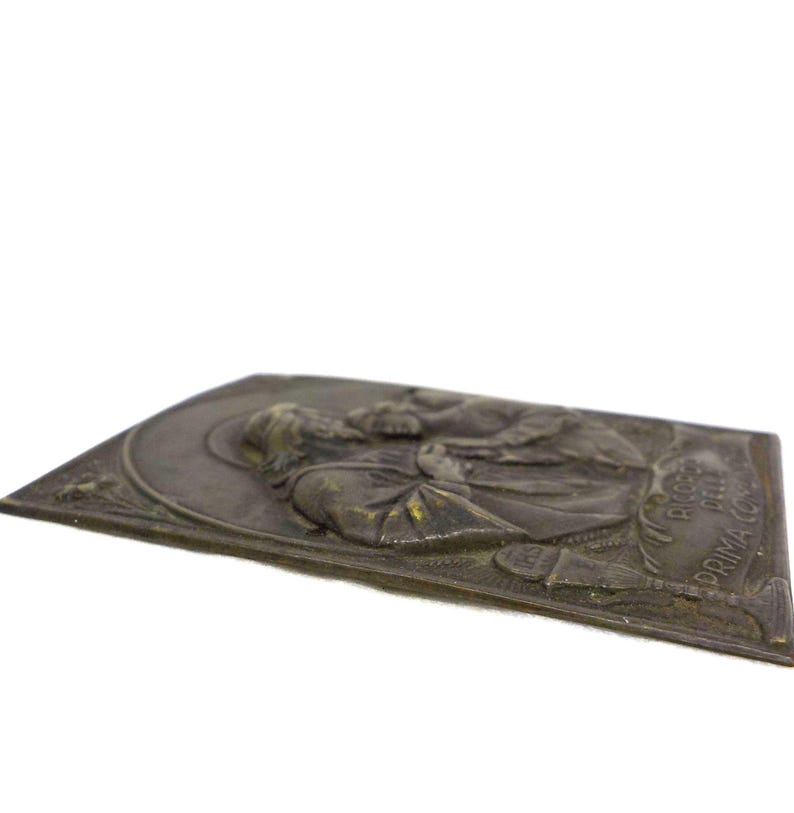 May include: A bronze-toned rectangular plaque with a relief of a religious figure. The figure is wearing a robe and has a halo. The plaque has text along the right edge. The plaque is slightly angled, showing its thickness.