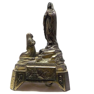 Peut inclure: Une figurine en métal doré représentant l'apparition de la Vierge Marie à Lourdes. La figurine présente une figure agenouillée d'une femme devant une figure debout de la Vierge Marie. La base de la figurine est inscrite du texte "Apparition de Lourdes".