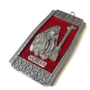May include: A rectangular, silver-toned religious plaque featuring a relief of a robed figure with a cross, set against a red velvet background. The plaque has a decorative border with floral patterns and the word "IZRITA".