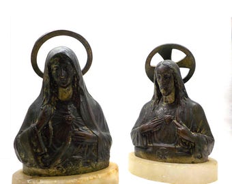 May include: Two bronze statues of religious figures, a woman and a man, with halos above their heads. Both figures are holding their hands over their hearts, and the statues are mounted on white marble bases.