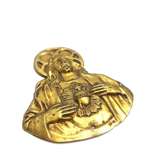 May include: A vintage, gold-toned metal religious pendant depicting a figure with a halo and a heart. The pendant has intricate details and a weathered appearance, suggesting age. The figure is holding their hands over their chest.