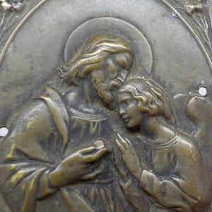 May include: Antique bronze religious plaque depicting a bearded figure with a halo, offering a small disc to a younger figure. The figures are in relief against an oval background, with floral accents and two small holes for hanging.