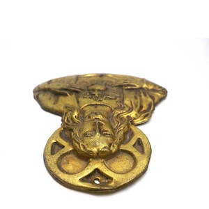 May include: A vintage brass-toned decorative piece featuring a detailed face with flowing hair and a circular base with a four-leaf clover design. The piece has an aged appearance and is set against a white background.