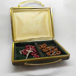 Könnte beinhalten: Eine offene, vintage-Stil Schmuckschatulle mit einem goldfarbenen Griff und Zierleisten. Im Inneren befindet sich ein grün mit Samt ausgekleidetes Fach mit zwei Rosenkränzen: einer mit roten Perlen und einem silbernen Kruzifix, und einer mit braunen Perlen.