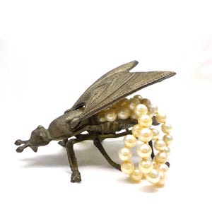 May include: A gold-toned metal fly figurine with its wings open, holding a strand of white pearls. The fly is a decorative trinket or jewellery holder.