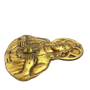 May include: A vintage, gold-toned metal religious pendant. The pendant features a detailed relief of a figure with long hair, holding a flower or sunburst. The figure is surrounded by a halo. The pendant has a hole for hanging.