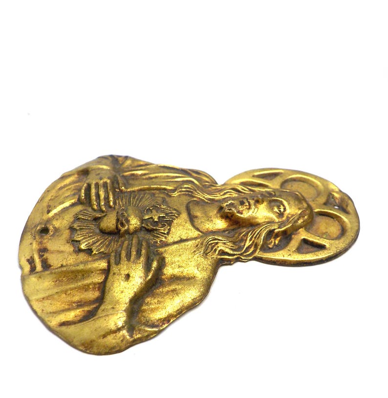 May include: A vintage, gold-toned metal religious pendant. The pendant features a detailed relief of a figure with long hair, holding a flower or sunburst. The figure is surrounded by a halo. The pendant has a hole for hanging.