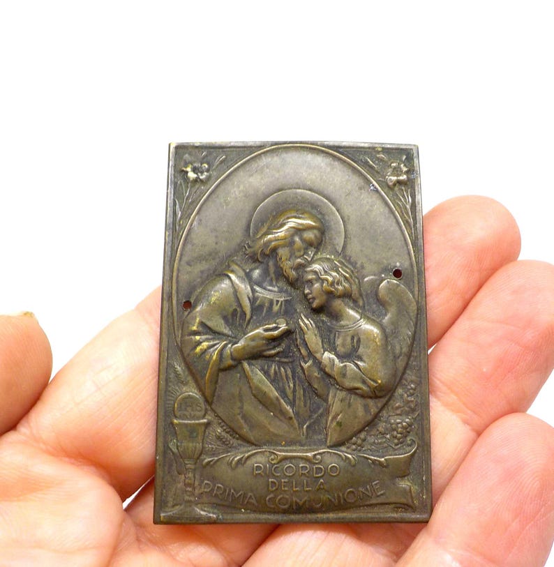 May include: A rectangular, antique-style religious plaque with a detailed relief. The plaque depicts two figures, one with a halo, and an angel. The text "RICORDO DELLA PRIMA COMUNIONE" is visible on a banner at the bottom. The plaque is held in a hand.