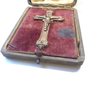 May include: An antique crucifix in a velvet-lined box. The crucifix is made of metal and depicts Jesus on the cross. The box is rectangular with a clasp closure and a faded, worn appearance. The velvet is a deep red color.