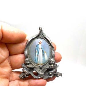 Könnte beinhalten: Vintage religiöser Anhänger mit einem ovalen Bild einer Frau in blauem Gewand, umrahmt von silberfarbenem Metall. Zwei Engel flankieren das Bild. Der Anhänger hat ein dekoratives, verziertes Design.