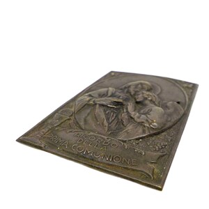 May include: A rectangular, antique-style metal plaque with an embossed religious scene. The plaque features a depiction of two figures, possibly a saint and a child, with the Italian text "RICORDO DELLA PRIMA COMUNIONE".