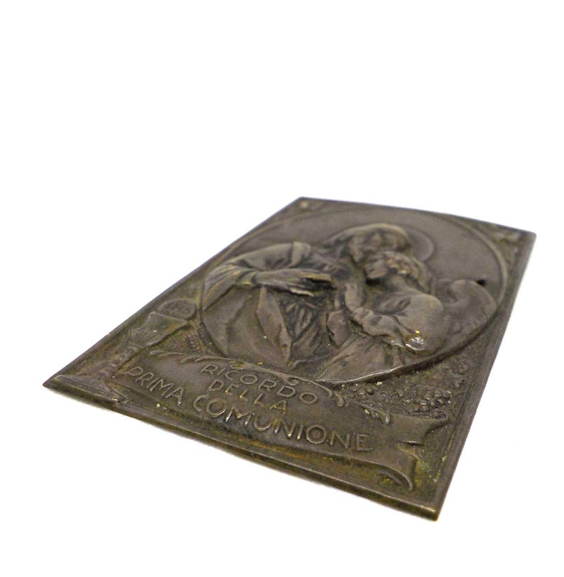 May include: A rectangular, antique-style metal plaque with an embossed religious scene. The plaque features a depiction of two figures, possibly a saint and a child, with the Italian text "RICORDO DELLA PRIMA COMUNIONE".