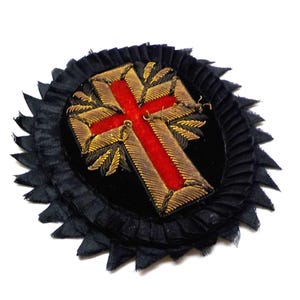 Pode incluir: Um emblema circular de tecido preto com uma cruz central de veludo vermelho delineada com bordado dourado metálico. O emblema é rodeado por uma borda de tecido preto plissado com um acabamento serrilhado e pontiagudo.